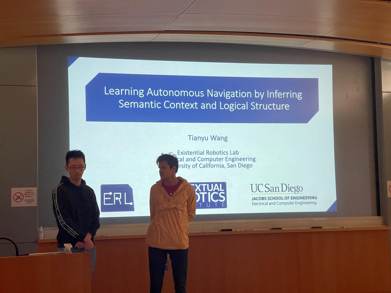 A RoboGrads member introduces the Feed the Intellect speaker in front of a projected slide reading "Learning Autonomous Navigation by Inferring Semantic Context and Logical Structure"